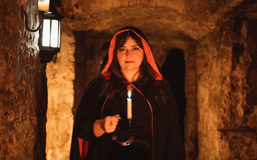 A guide in a hooded cloak holding a candle in the vaults
